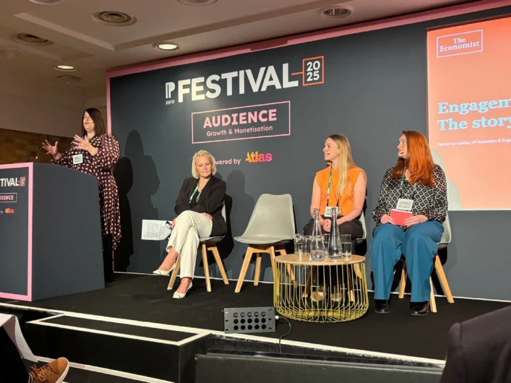 Tamsin Larcombe, VP retention at The Economist, speaking at the PPA Festival standing at a podium on stage. Also on stage are three other women seated with head mics on listening to Larcombe