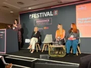 Tamsin Larcombe, VP retention at The Economist, speaking at the PPA Festival standing at a podium on stage. Also on stage are three other women seated with head mics on listening to Larcombe