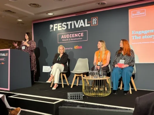 Tamsin Larcombe, VP retention at The Economist, speaking at the PPA Festival standing at a podium on stage. Also on stage are three other women seated with head mics on listening to Larcombe