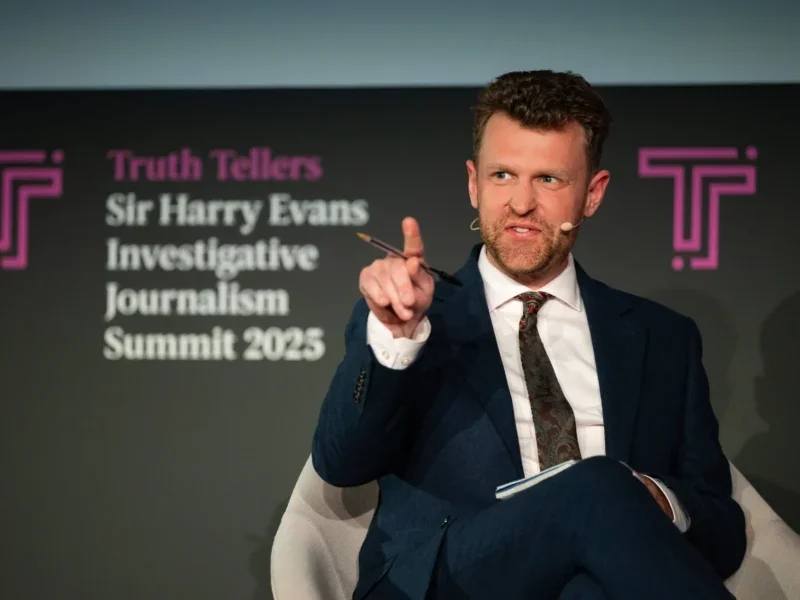 New Statesman editor-in-chief Tom McTague appears on stage at the Sir Harry Evans Investigative Journalism Summit in London in 2025. McTague, dressed in a dark suit and with a mic affixed to his cheek, is seen raising a finger and looking off camera toward the crowd.