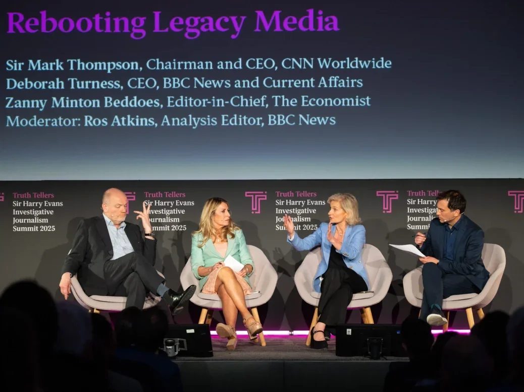 Left to right: CNN chief executive Mark Thompson, BBC News chief executive Deborah Turness, The Economist editor-in-chief Zanny Minton Beddoes and BBC analysis editor Ros Atkins. The three executives are visibly mid-disagreement: Minton Beddoes is gesticulating with her hands apart as though making a firm point, Turness looks taken aback and Thompson is leaned back casting a single hand up with the fingers splayed as though to suggest he is either taking the point or thinks it's immaterial.
