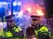 Two police officers at nighttime with riot shields in front of them and fire behind them
