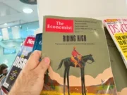 Man holds The Economist magazine in print in a shop with other magazines on the newsstand behind it