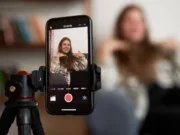 Woman sitting on a sofa as seen through a phone screen which is attached to a tripod. The woman in real life is seen blurred to the side of the screen