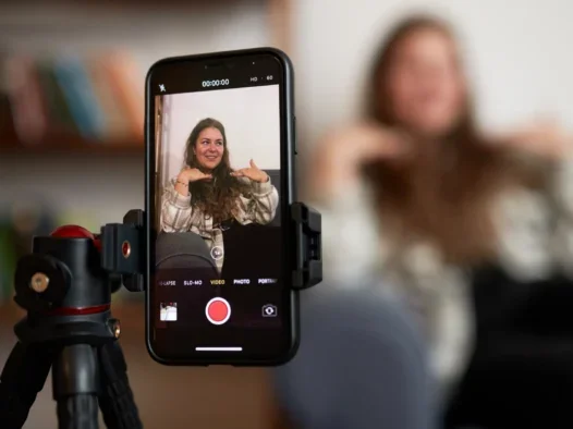 Woman sitting on a sofa as seen through a phone screen which is attached to a tripod. The woman in real life is seen blurred to the side of the screen