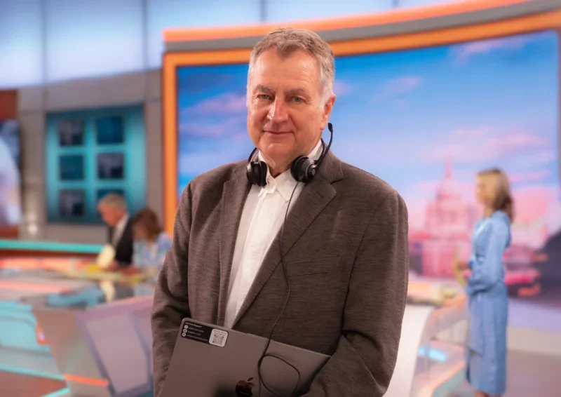 Headshot of Long-serving Good Morning Britain editor Neil Thompson standing in the TV studio with headphones around his neck, wearing a suit with no tie and holding a laptop