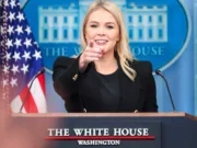 White House Press Secretary Karoline Leavitt standing at White House branded lectern pointing at someone behind the camera. She has a White House seal on the wall behind her and an American flag