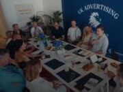 About 12 people sitting around a large rectangular table either listening or speaking, clearly mid-discussion. Wall behind them displays the UK Advertising logo