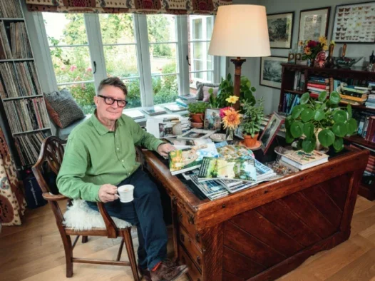 Mark Hedges, Country Life editor-in-chief sitting at a wooden desk which features piles of Country Life magazines, holding a cup of tea. Large window behind him looks onto a large garden