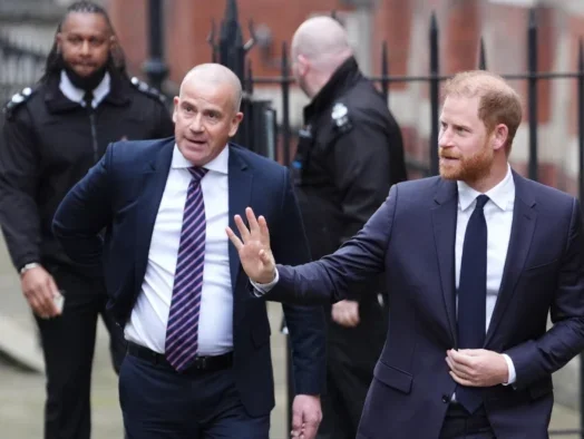 Prince Harry walking outside a court building waving at people out of shot, with one man in a suit just behind him and two people in court security uniforms further behind