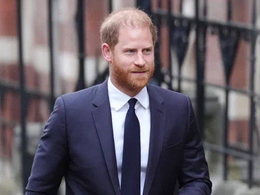 Prince Harry smiling looking out of shot wearing a suit and tie, walking with railings behind him in the street
