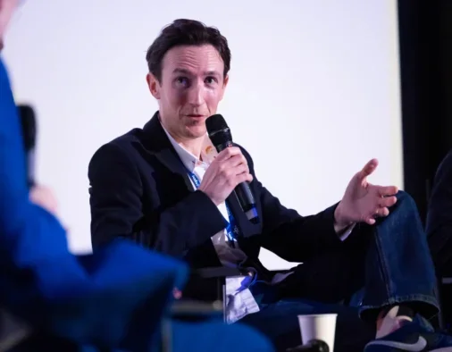 Captify global data science and insights director Sam Coates speaking on stage, holding microphone with one hand and gesturing with the other