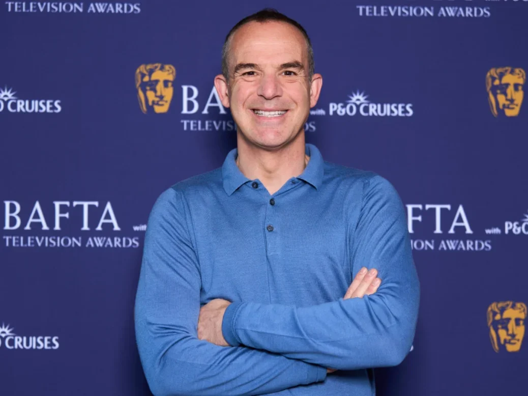 Martin Lewis stands smiling with arms crossed wearing blue shirt in front of background featuring BAFTA and P&O Cruises logos