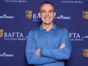 Martin Lewis stands smiling with arms crossed wearing blue shirt in front of background featuring BAFTA and P&O Cruises logos
