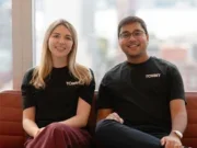 Tollbit co-founders Olivia Joslin and Toshit Panigrahi sitting side by side on a sofa smiling at the camera, both wearing black Tollbit branded t-shirts