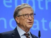 Bill Gates wearing a suit and tie, speaking into a microphone with a blue stage background behind him