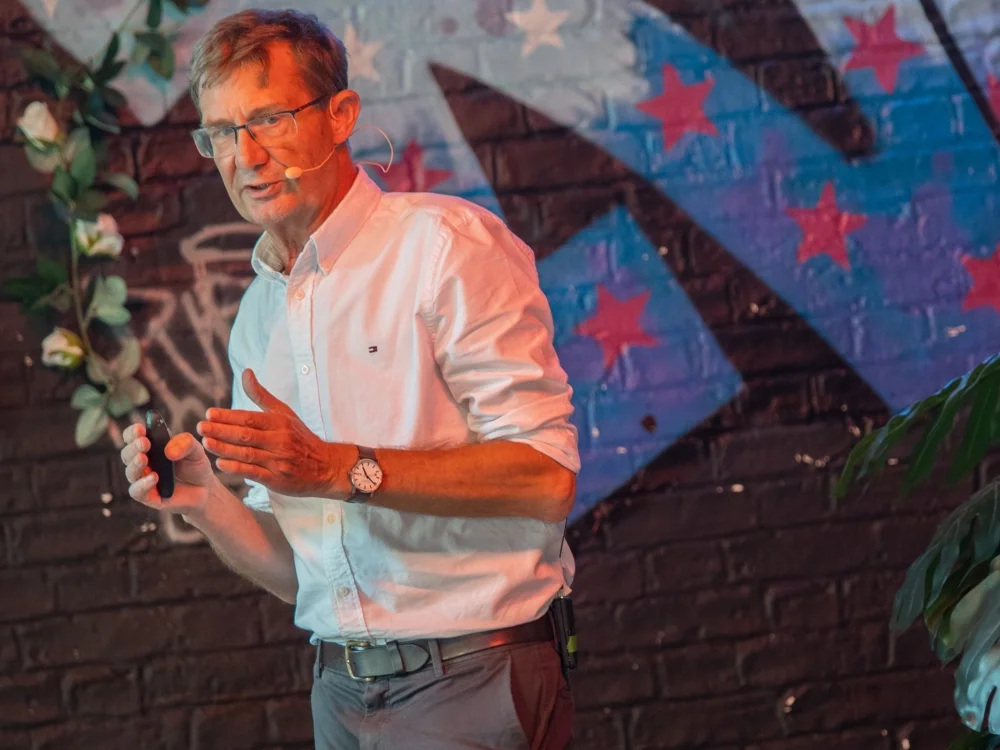 Nic Newman standing up speaking and gesturing with hands with brightly coloured wall behind him