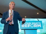 Nigel Farage standing next to Reform-branded lectern holding glass of champagne