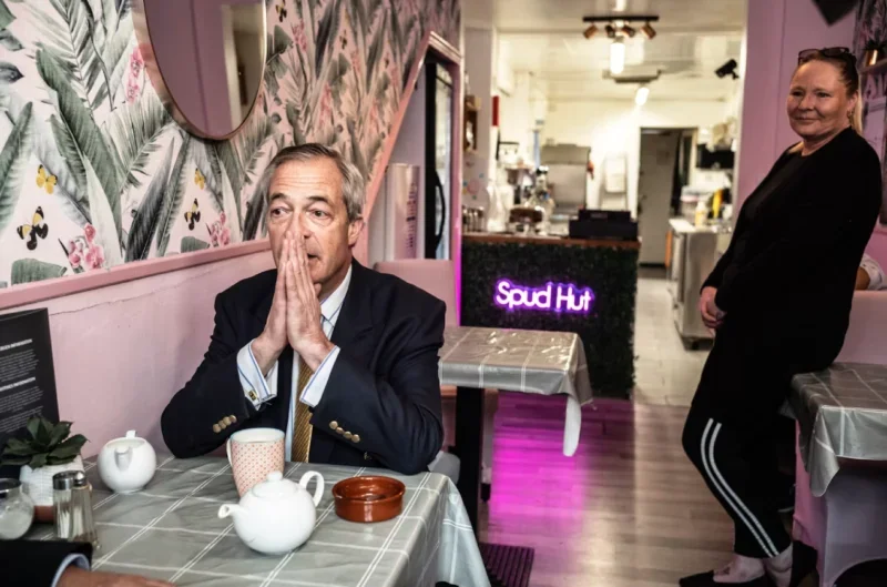 Image of Nigel Farage alone at a cafe table with hands pressed together in front of his face